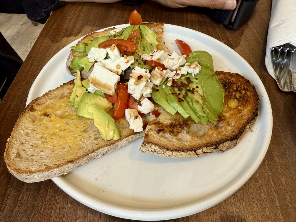Sliced green avocade, with red cherry tomatoes, feta cheese and chili flakes on three slices of toasted sourdough bread
