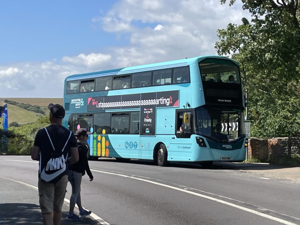 Turquoise double decker bus traveling along a rural road with rolling hills, trees, and a partly cloudy sky in the background. Two pedestrians wait at the roadside as the bus passes, illustrating a public transportation option for travelers