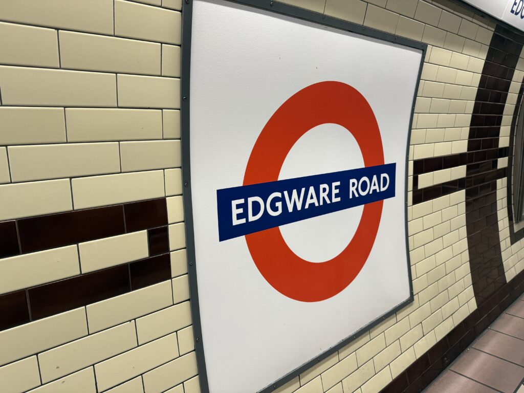 London Underground station sign displaying "EDGWARE ROAD" on a white panel with the iconic red roundel, mounted against a cream tiled wall with dark brown accents. The sign marks a Tube station that is part of London's public transit network, highlighting one of the easiest ways to get around London.
