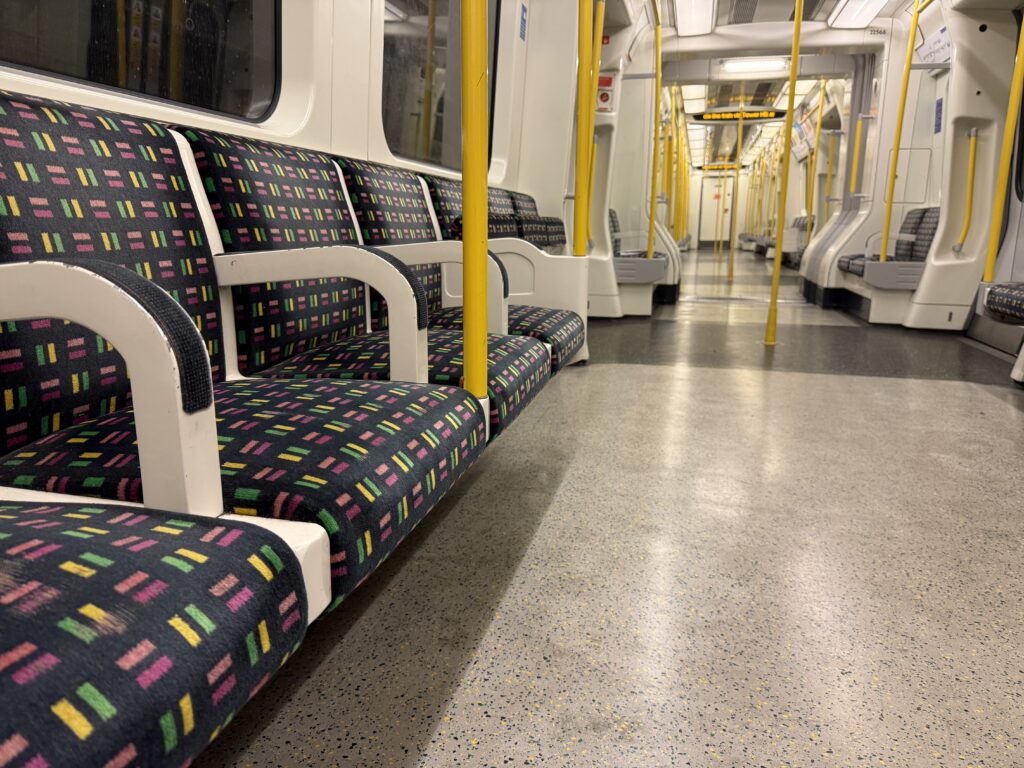 Interior of a London Underground train with patterned seats, bright yellow grab poles, and a wide central aisle stretching through an empty carriage. The clean, modern train highlights one of the fastest and most convenient ways to get around